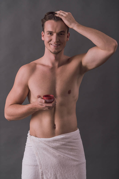 Beau jeune homme avec torse nu, enveloppé dans une serviette utilise un gel capillaire, regardant la caméra et souriant, sur fond gris
 - Photo, image