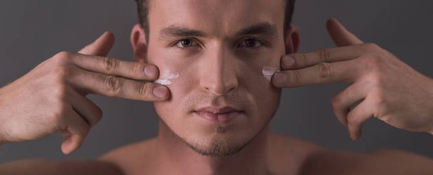 Handsome young man is applying face cream and looking at camera, on gray background - Foto, Bild