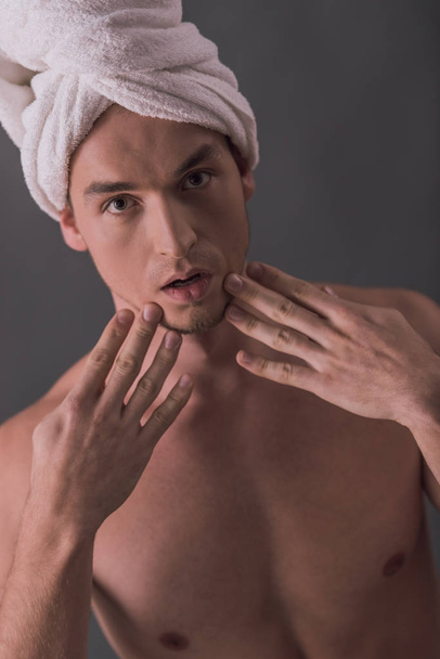 Όμορφος νεαρός άνδρας με γυμνό κορμό και την πετσέτα στο κεφάλι του αγγίζει το πρόσωπό του, σε γκρίζο φόντο - Φωτογραφία, εικόνα