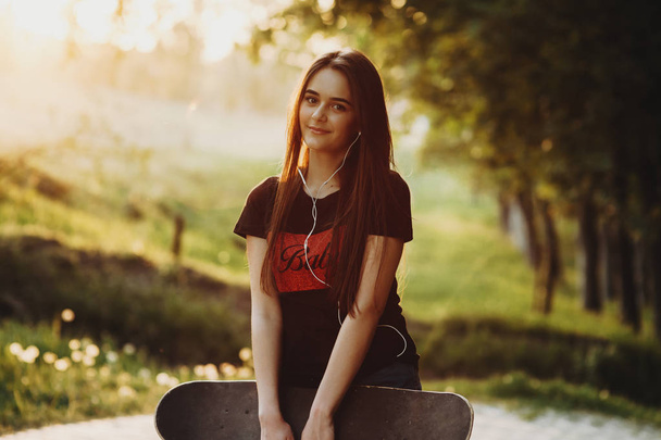 Portrait d'une charmante jeune fille aux cheveux longs assise dans un parc contre le coucher du soleil avec une planche à roulettes sur les mains regardant dans la caméra souriant tout en écoutant de la musique dans les écouteurs
. - Photo, image