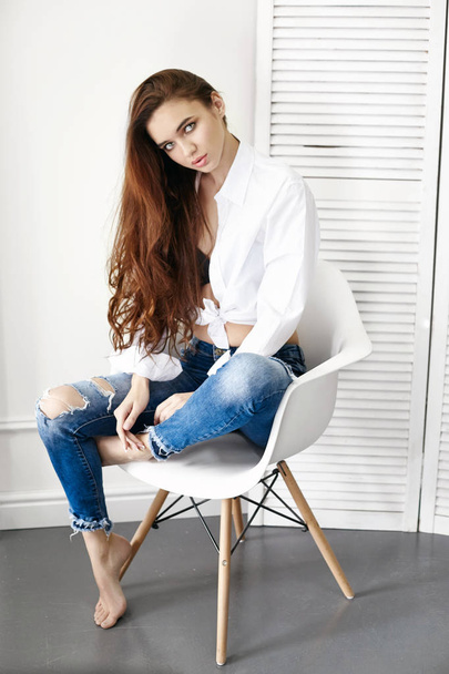 Sexy beautiful girl in jeans white shirt sitting on a chair. Gorgeous long hair and charming eyes young woman. Sexy Business lady is young and confident. Girl sitting on chair at home, naked body - Fotó, kép