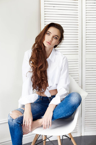 Sexy beautiful girl in jeans white shirt sitting on a chair. Gorgeous long hair and charming eyes young woman. Sexy Business lady is young and confident. Girl sitting on chair at home, naked body - Фото, изображение