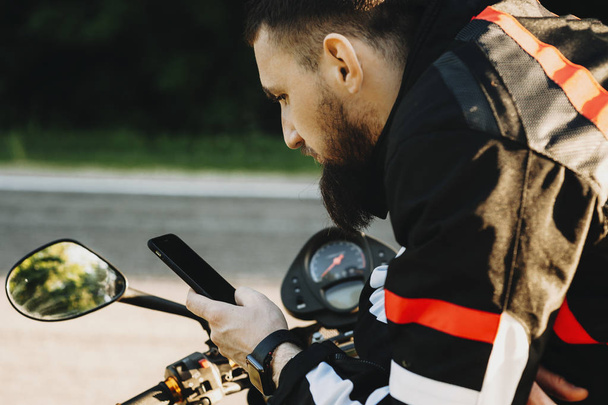 Красивый бородатый путешественник опирается на свой велосипед, глядя в смартфон, чтобы найти маршрут рядом с дорогой
. - Фото, изображение