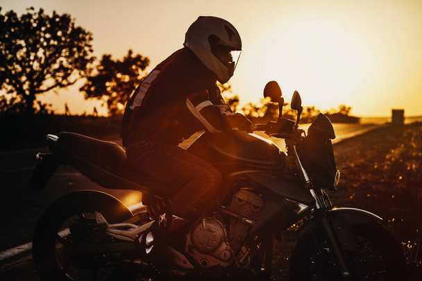 Vista lateral de um jovem motociclista caucasiano sentado em sua bicicleta e descansando em seu tempo de férias contra o pôr do sol
. - Foto, Imagem