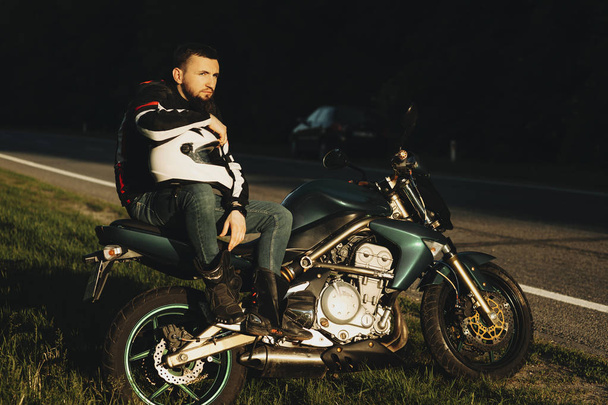 Full length portrait of a handsome serious biker sitting on his motorcycle with a hand on his helmet and looking away near the road against the forest. - Foto, Imagen