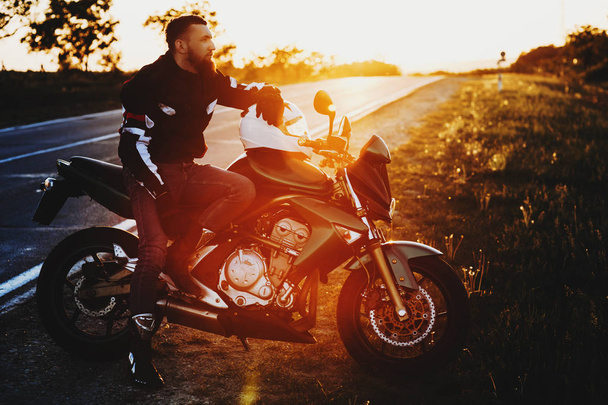 Retrato completo de un hombre caucásico joven impresionante sentado en su motocicleta mientras descansa en su viaje de vacaciones cerca de la carretera contra la puesta del sol
. - Foto, imagen