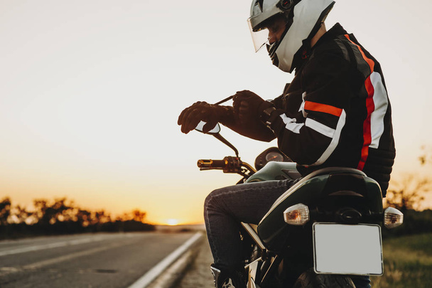 Vista lateral de un hombre caucásico sentado en la motocicleta y poniendo sus guantes preparándose para comenzar su aventura en motocicleta contra el atardecer
. - Foto, Imagen