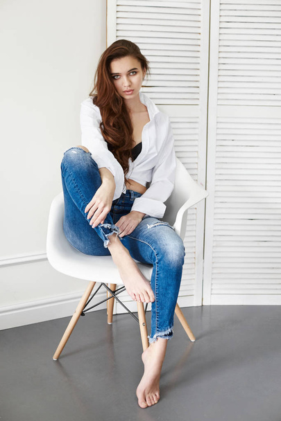 Sexy beautiful girl in jeans white shirt sitting on a chair. Gorgeous long hair and charming eyes young woman. Sexy Business lady is young and confident. Girl sitting on chair at home, naked body - Foto, Imagem