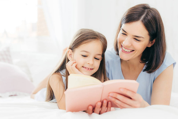Die hübsche Mutter und ihre kleine Tochter lesen im Schlafzimmer interessante Bücher, liegen morgens auf einem bequemen Bett. schöne Mutter liest Märchen für ihr kleines weibliches Kind, das sehr neugierig ist - Foto, Bild