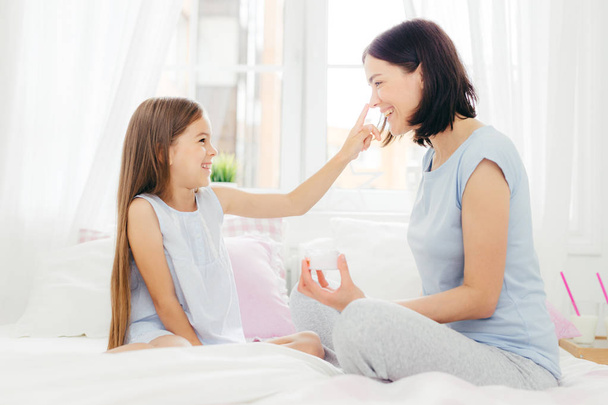 Pretty small female child has fun together with her mother, applies beauty cream on her nose, sit together on comfortable bed, pose in spacious light bedroom. People, happiness and family concept - Foto, Imagen