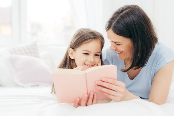 Indoor shot of pleasant looking female child with curious expression, reads interesting book together with her affectionate mother, views colourful pictures, lie on bed in spacious bedroom. Childhood - Photo, Image
