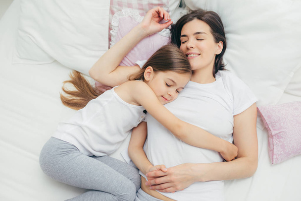 Top view of cheerful mother sleeps on white bed near her daughter who embraces mum with great love, shows good attitude, have pleasant dreams and nice rest, wears pyjamas. People, sleeping concept - Foto, Bild