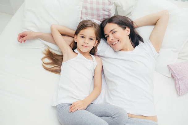 Innenaufnahme von fröhlicher Mutter und Tochter, die sich umarmen, wie sie auf weißen Bettwäschen liegen, morgens aufwachen, Wochenende haben, im Schlafzimmer sein, zufriedene Mienen haben. Menschen, Lebensstil-Konzept - Foto, Bild