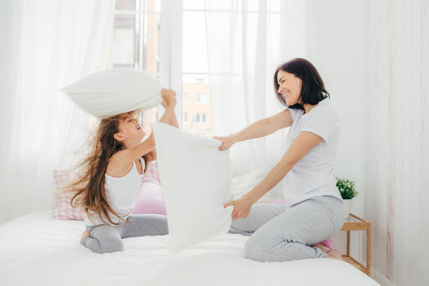 Indoor shot of pleasant looking European female has joy together with her small daughter, have pillow fight in bedroom, pose in cozy spacious light room, being in high spirit. People and fun concept - Foto, imagen