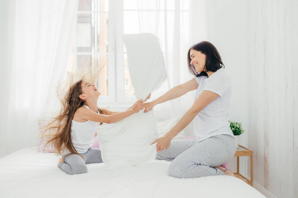 Parenthood and family concept. Attractive female mother with positive smile, spends free time with her daughter, have pillow fight, pose against bedroom interior, enjoys leisure time during weekend - Photo, image