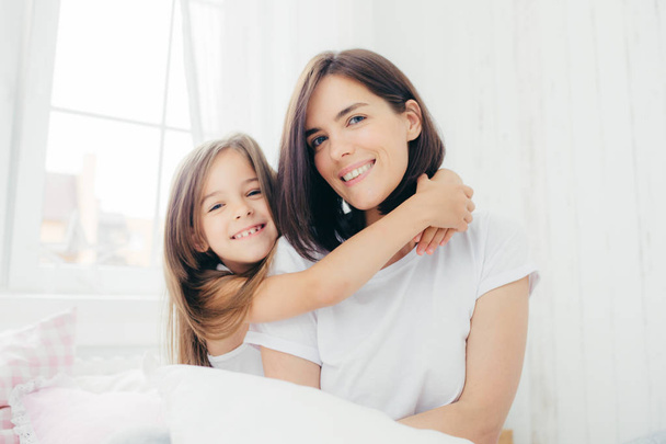 Indoor shot of good looking brunette mother with gentle smile and her small daughter gives hug, enjoy domestic atmosphere, pose against spacious white bedroom interior. Motherhood and family concept - Photo, Image