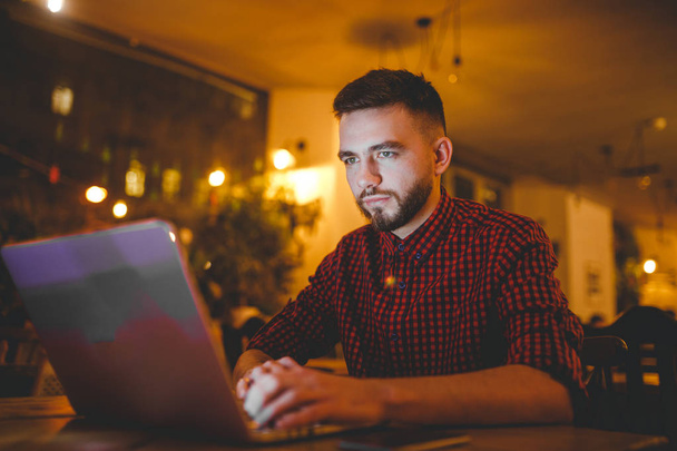 Hinter einem grauen Laptop sitzt an einem Holztisch ein junger gutaussehender kaukasischer Mann mit Bart und zahmem Lächeln im rot karierten Hemd. Hände auf der Tastatur. Abends im Café - Foto, Bild