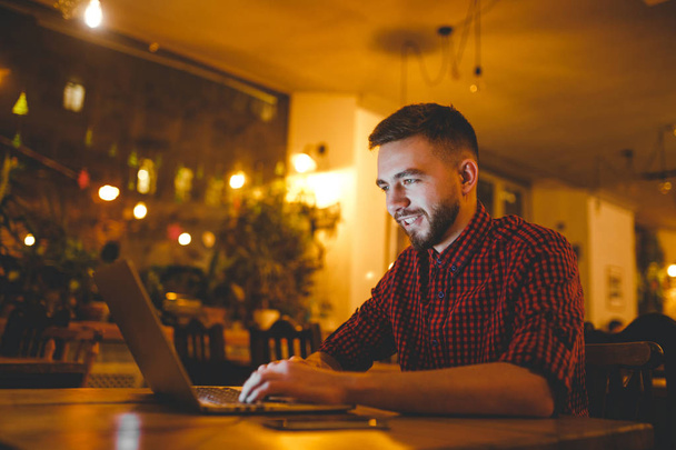 Egy fiatal csinos kaukázusi Férfi szakáll és toothy mosoly, egy piros kockás inget mögött ül egy fa asztal szürke laptop dolgozik. Kezek-on billentyűzet. A kávézó este - Fotó, kép