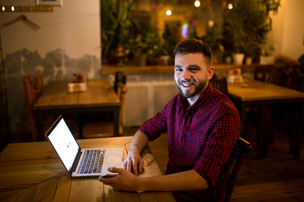 Młody przystojny kaukaski mężczyzna z brodą i Uśmiech zębiczny w czerwone kraciaste koszule działa za laptopa siedząc na drewnianym stole. Używa trzyma w ręku telefon komórkowy. Wieczorem w kawiarni - Zdjęcie, obraz