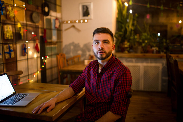 Porträt eines jungen gutaussehenden kaukasischen Mannes mit Bart und zahmem Lächeln in einem rot karierten Hemd, der neben einem Laptop an einem Holztisch sitzt. Abends im Café - Foto, Bild