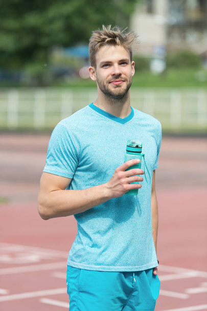 Man with athletic appearance holds bottle with water. Athlete drink water after training at stadium on sunny day. Sport and healthy lifestyle concept. Man athlete in sporty clothes training outdoor - Fotó, kép