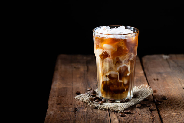 背の高いグラスに注がれるクリームと古い素朴な木製のテーブルの上のコーヒー豆のアイス コーヒー。コピー スペースと暗い背景の木に冷たい夏の飲み物. - 写真・画像