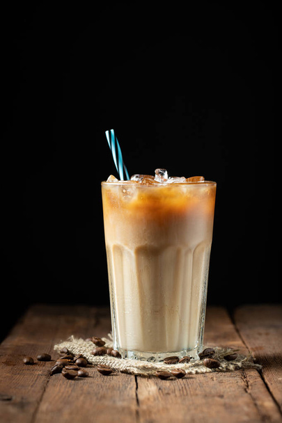 Ice coffee in a tall glass with cream poured over and coffee beans on a old rustic wooden table. Cold summer drink on a dark wooden background with copy space. - Photo, Image