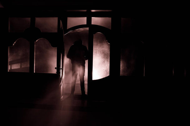 閉じたガラスのドアを通ってドアの上に未知の影の姿のシルエット。夜の窓の前に人間のシルエット。怖いシーンハロウィンの概念のぼやけたシルエットのマニアック. - 写真・画像