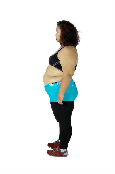 Side view of obese woman looks pensive while wearing sportswear and standing in the studio, isolated on white background - Photo, image
