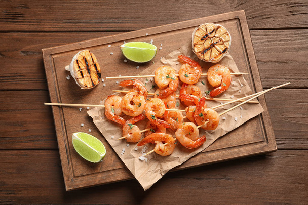 Board with delicious fried shrimp skewers, lime and garlic on wooden background, top view - Foto, imagen