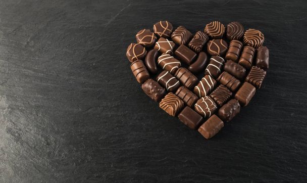 Όμορφη δημιουργική σοκολάτας καρδιά σχήμα κατασκευασμένο με διάφορους τύπους πραλίνες πάνω από ένα μαύρο φόντο πέτρα. Γλυκά σοκολάτες με μαύρο φόντο - Φωτογραφία, εικόνα