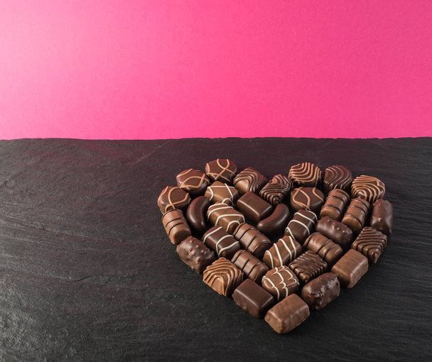 Όμορφη δημιουργική σοκολάτας καρδιά σχήμα κατασκευασμένο με διάφορους τύπους πραλίνες πάνω από ένα μαύρο φόντο πέτρα. Γλυκά σοκολάτες με φόντο το ροζ - Φωτογραφία, εικόνα