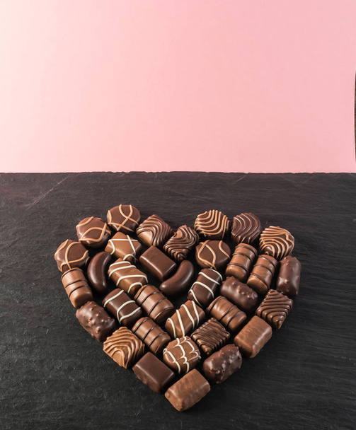 Όμορφη δημιουργική σοκολάτας καρδιά σχήμα κατασκευασμένο με διάφορους τύπους πραλίνες πάνω από ένα μαύρο φόντο πέτρα. Γλυκά σοκολάτες με φόντο το ροζ - Φωτογραφία, εικόνα