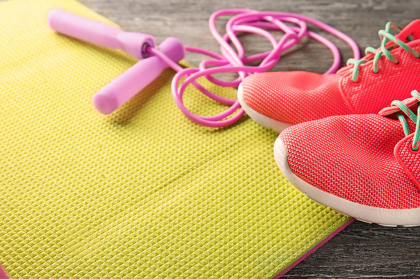 Yoga mat with jump rope and sneakers on wooden background, closeup - Fotó, kép