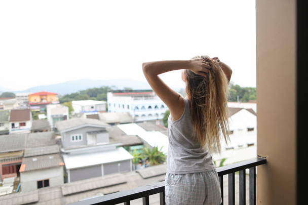 Young blonde girl wearing summer pajamas standing on balcony and looking at buildings with raised hands. - Photo, Image