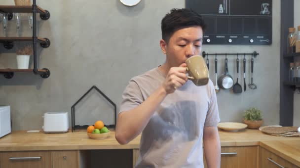 Happy handsome asian young man drinking a cup of coffee. Attractive asian man eating apple while staying in the kitchen. Male using time at home. - Video