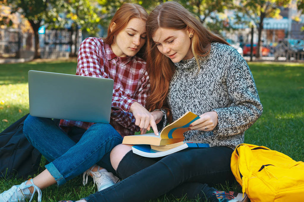 Attractive smiling students studying outdoors on campus at the university. Beautiful college girls with reddish hair sit on campus grass with crossed legs using laptop pointing with finger in books.  - 写真・画像