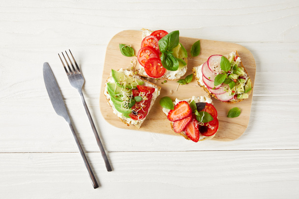 top view of various sandwiches on cutting board with cutlery on white wooden surface - Foto, immagini