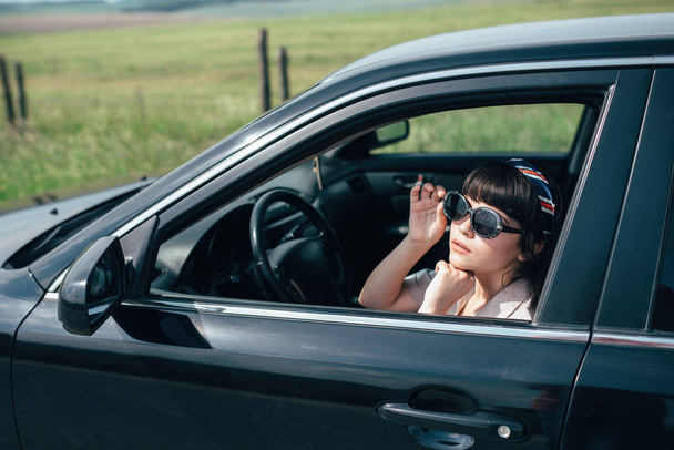 Güzel genç mavi gökyüzü ve alan arka plan üzerinde siyah bir araba direksiyonuna oturan kız. - Fotoğraf, Görsel