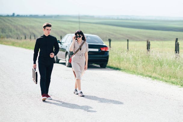 Couple de jeune fille et garçon tenant des pourparlers près de voiture noire sur fond de ciel bleu et champ
. - Photo, image