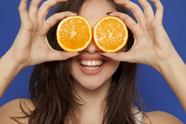 Happy young woman posing with slices of orange on her face on a blue background - Photo, Image