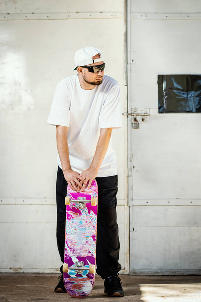 Mann mit Mütze hält gebrauchtes Skateboard im Sommer in einem Betonskatepark in den Händen - Foto, Bild