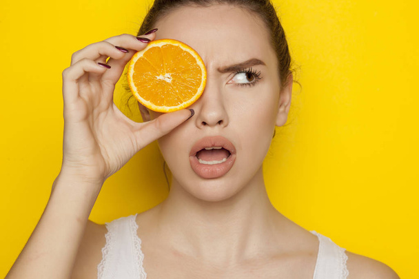 Surprised young woman posing with slice of orange on her face on a yellow background - Photo, Image