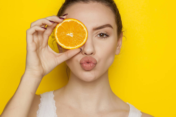 Молода жінка позує зі скибочкою апельсина на її обличчі на жовтому фоні
 - Фото, зображення