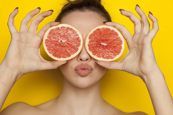 Happy sexy woman posing with slices of red grapefruit on her face on a yellow background - Φωτογραφία, εικόνα