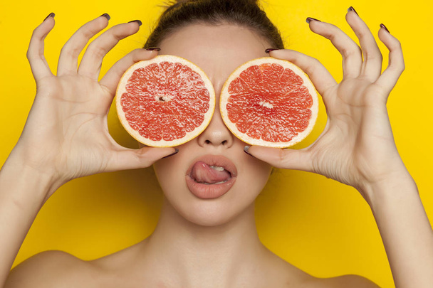 Mladá sexy žena pózuje s plátky červeného grapefruitu na její obličej na žlutém podkladu - Fotografie, Obrázek