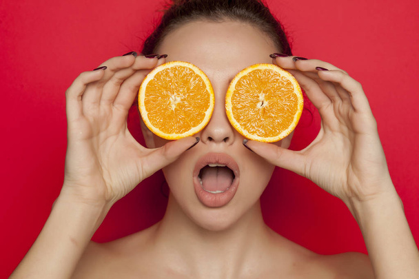 Удивлённая девушка позирует с кусками апельсинов на лице на красном фоне.
 - Фото, изображение