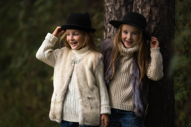 Two happy girls are equally dressed: in fur vests and hats in the forest. Little girlfriends in park. Children Friendship Together Smiling Happiness Concept - Fotoğraf, Görsel