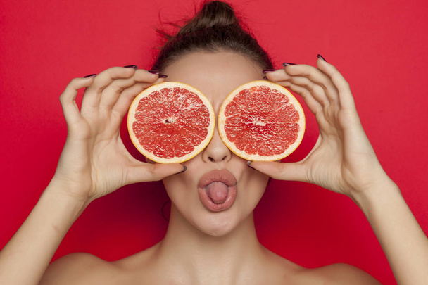Happy sexy woman posing with slices of red grapefruit on her face on a red background - Photo, image