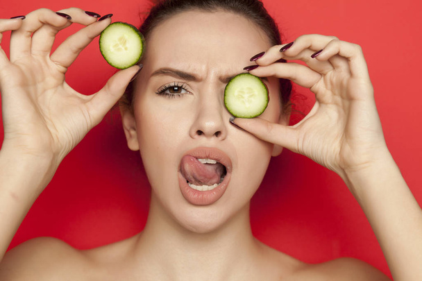Sexy young woman posing with slices of cucumber on her face on a red background - Photo, Image